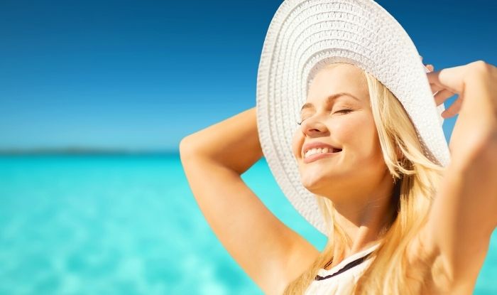 Mujer con sombrero en la playa sonriendo al sol con los ojos cerrados.